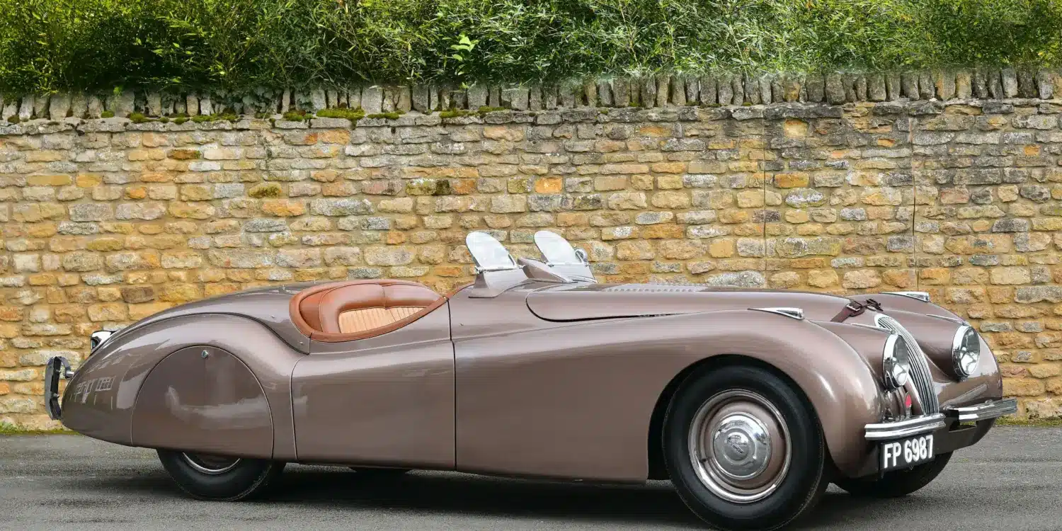 Seitenansicht eines braunen Jaguar XK120 Roadster von 1950 vor einer Steinmauer