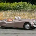 Seitenansicht eines braunen Jaguar XK120 Roadster von 1950 vor einer Steinmauer
