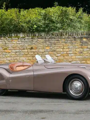 Seitenansicht eines braunen Jaguar XK120 Roadster von 1950 vor einer Steinmauer