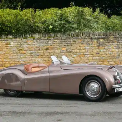 Seitenansicht eines braunen Jaguar XK120 Roadster von 1950 vor einer Steinmauer