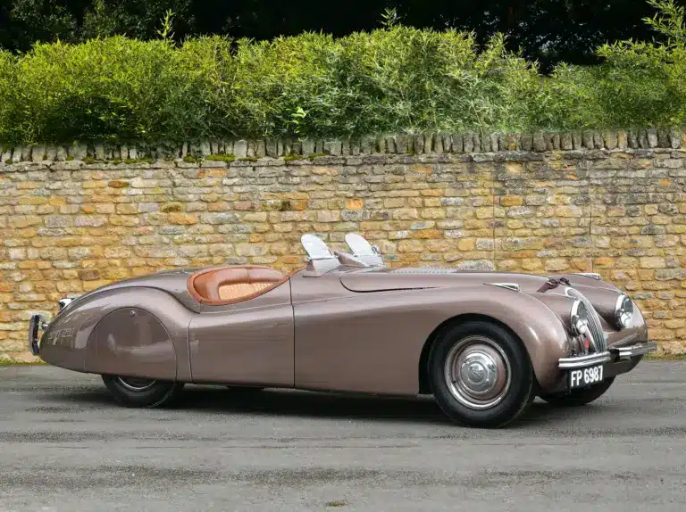 Seitenansicht eines braunen Jaguar XK120 Roadster von 1950 vor einer Steinmauer