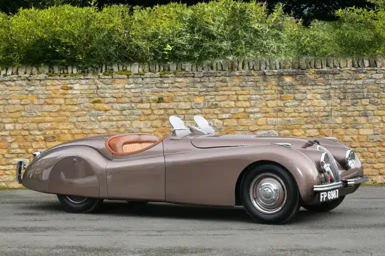 Seitenansicht eines braunen Jaguar XK120 Roadster von 1950 vor einer Steinmauer