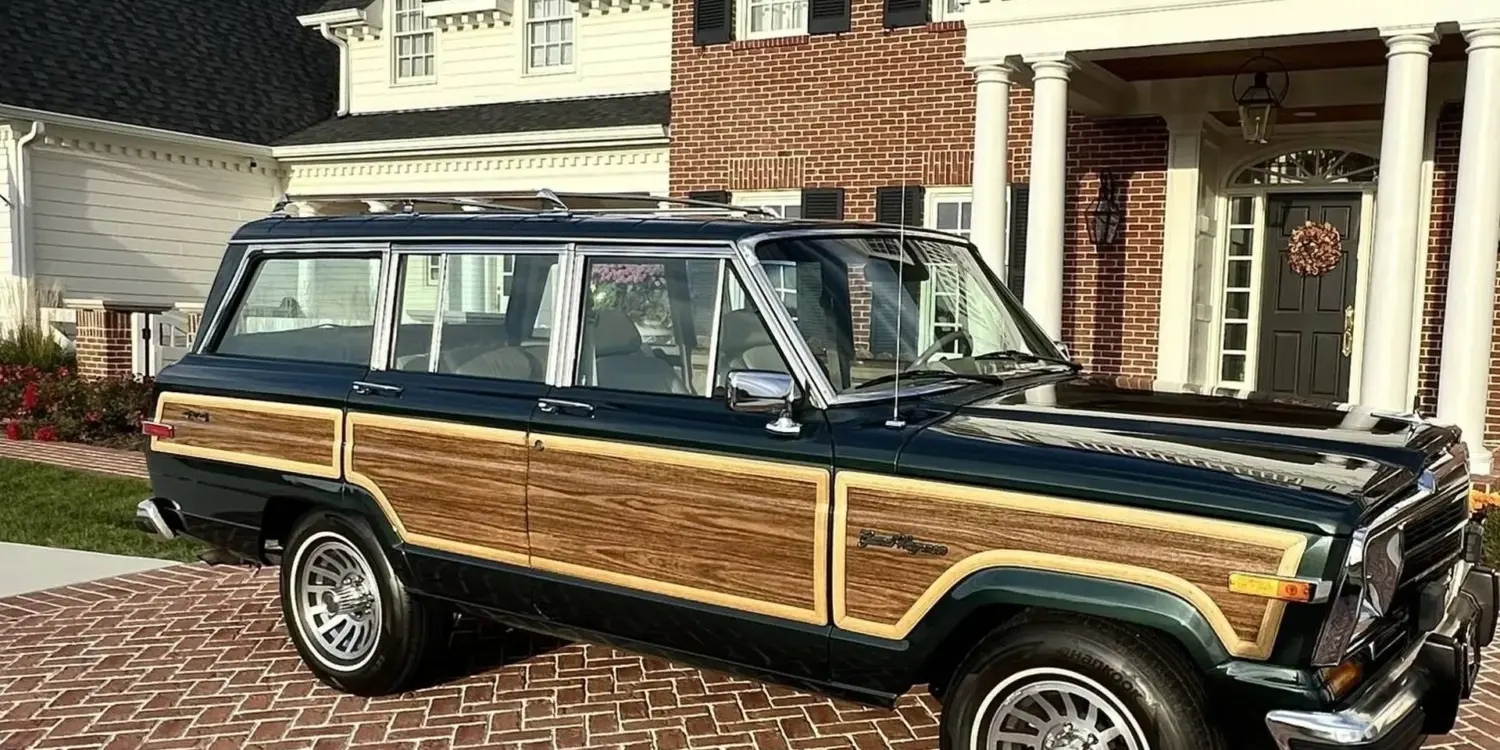 Ein 1991 Jeep Grand Wagoneer mit Holzdekor-Elementen steht auf einem gepflasterten Hof vor einem großen, weißen Anwesen.