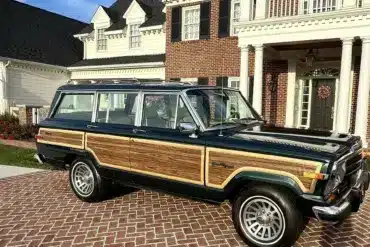 Ein 1991 Jeep Grand Wagoneer mit Holzdekor-Elementen steht auf einem gepflasterten Hof vor einem großen, weißen Anwesen.