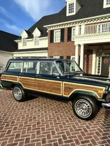Ein 1991 Jeep Grand Wagoneer mit Holzdekor-Elementen steht auf einem gepflasterten Hof vor einem großen, weißen Anwesen.