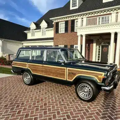 Ein 1991 Jeep Grand Wagoneer mit Holzdekor-Elementen steht auf einem gepflasterten Hof vor einem großen, weißen Anwesen.