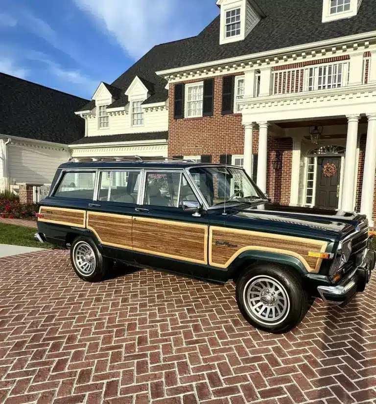 Ein 1991 Jeep Grand Wagoneer mit Holzdekor-Elementen steht auf einem gepflasterten Hof vor einem großen, weißen Anwesen.