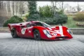 Roter Lola T70 Mk3B Rennwagen mit Startnummer 19, fotografiert von schräg vorne auf Pflastersteinen.