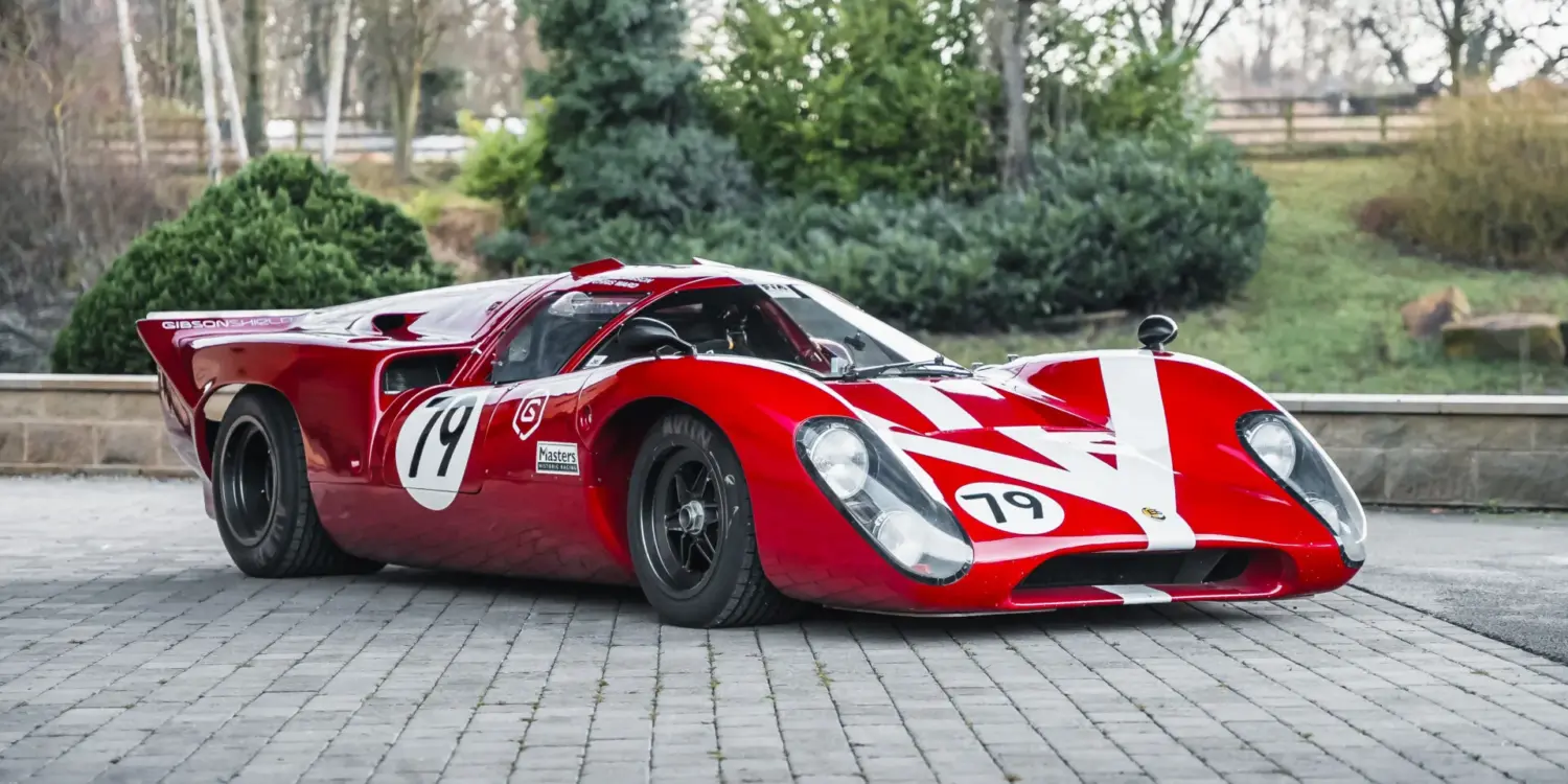 Roter Lola T70 Mk3B Rennwagen mit Startnummer 19, fotografiert von schräg vorne auf Pflastersteinen.