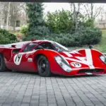 Roter Lola T70 Mk3B Rennwagen mit Startnummer 19, fotografiert von schräg vorne auf Pflastersteinen.