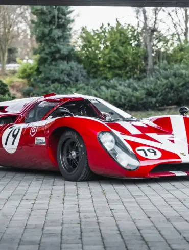 Roter Lola T70 Mk3B Rennwagen mit Startnummer 19, fotografiert von schräg vorne auf Pflastersteinen.