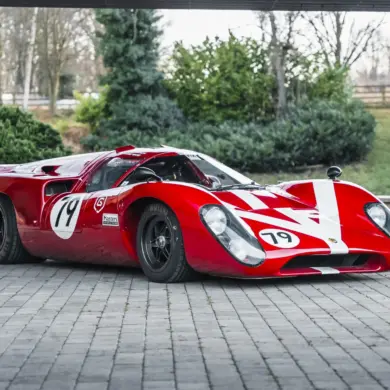 Roter Lola T70 Mk3B Rennwagen mit Startnummer 19, fotografiert von schräg vorne auf Pflastersteinen.
