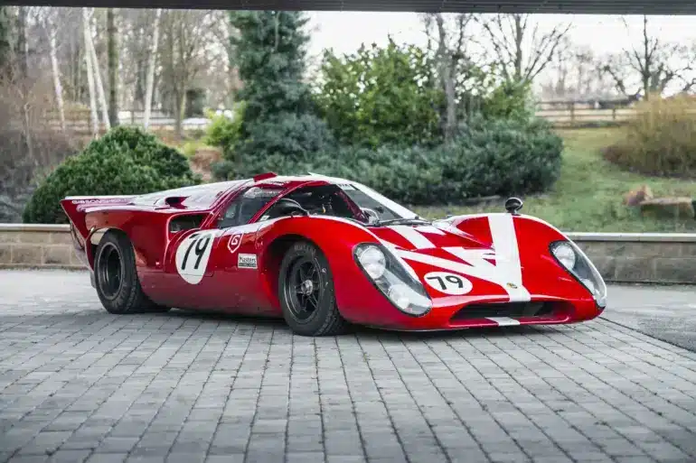 Roter Lola T70 Mk3B Rennwagen mit Startnummer 19, fotografiert von schräg vorne auf Pflastersteinen.