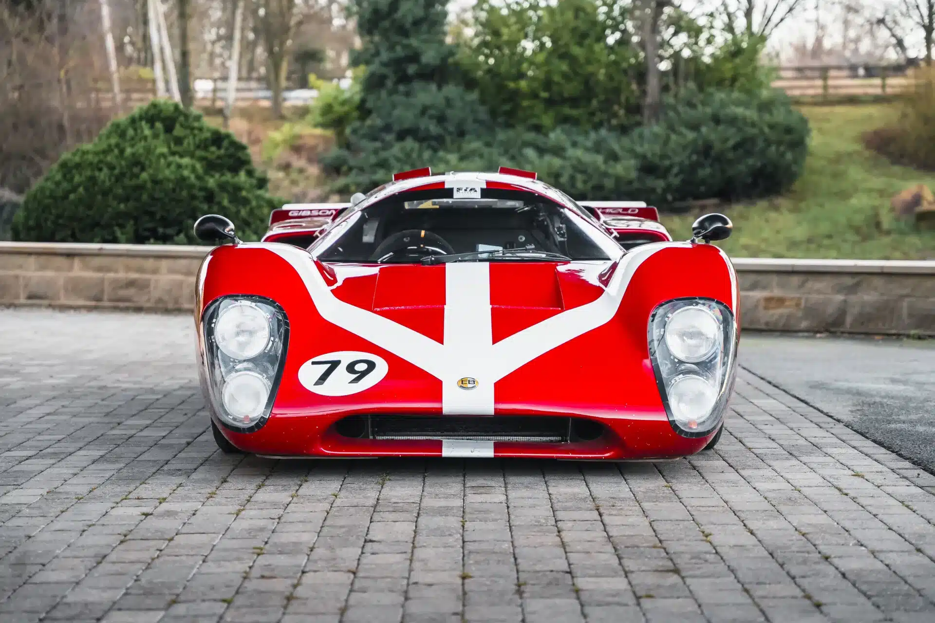 Frontansicht eines roten Lola T70 Mk3B mit weißem Streifen und Startnummer 79, stehend auf Pflastersteinen.