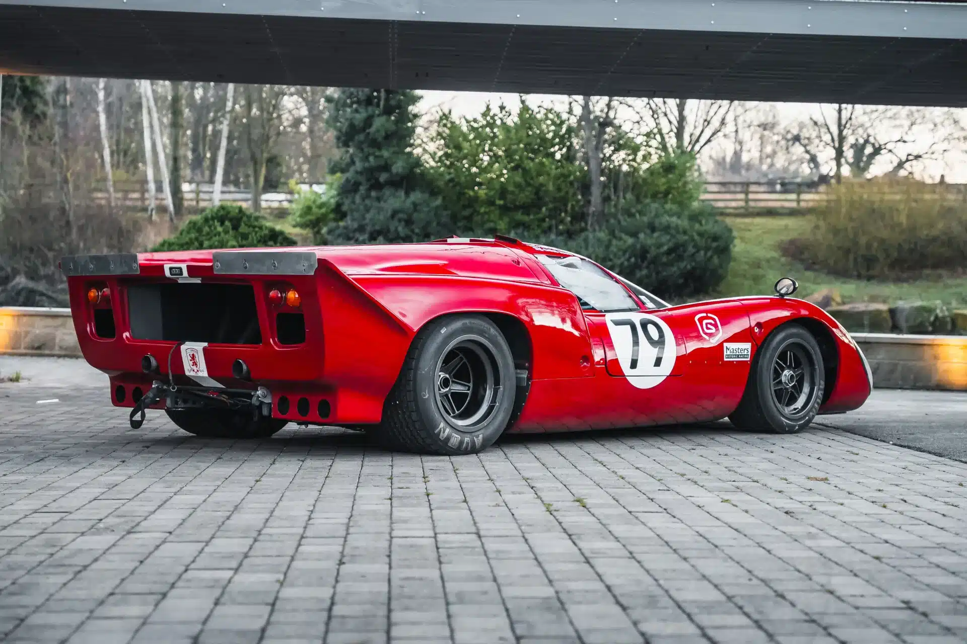 Seiten- und Heckansicht eines roten Lola T70 Mk3B Rennwagens mit Startnummer 79, stehend auf einem Pflastersteinboden.