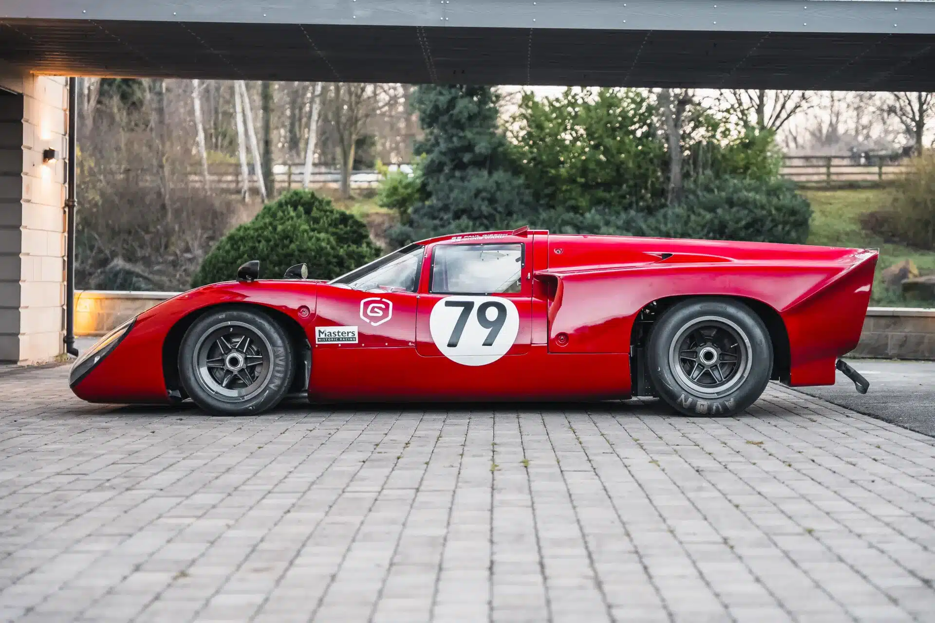 Lola T70 Mk3B in Rot, Seitenansicht, steht auf Pflastersteinen vor einer Garage.