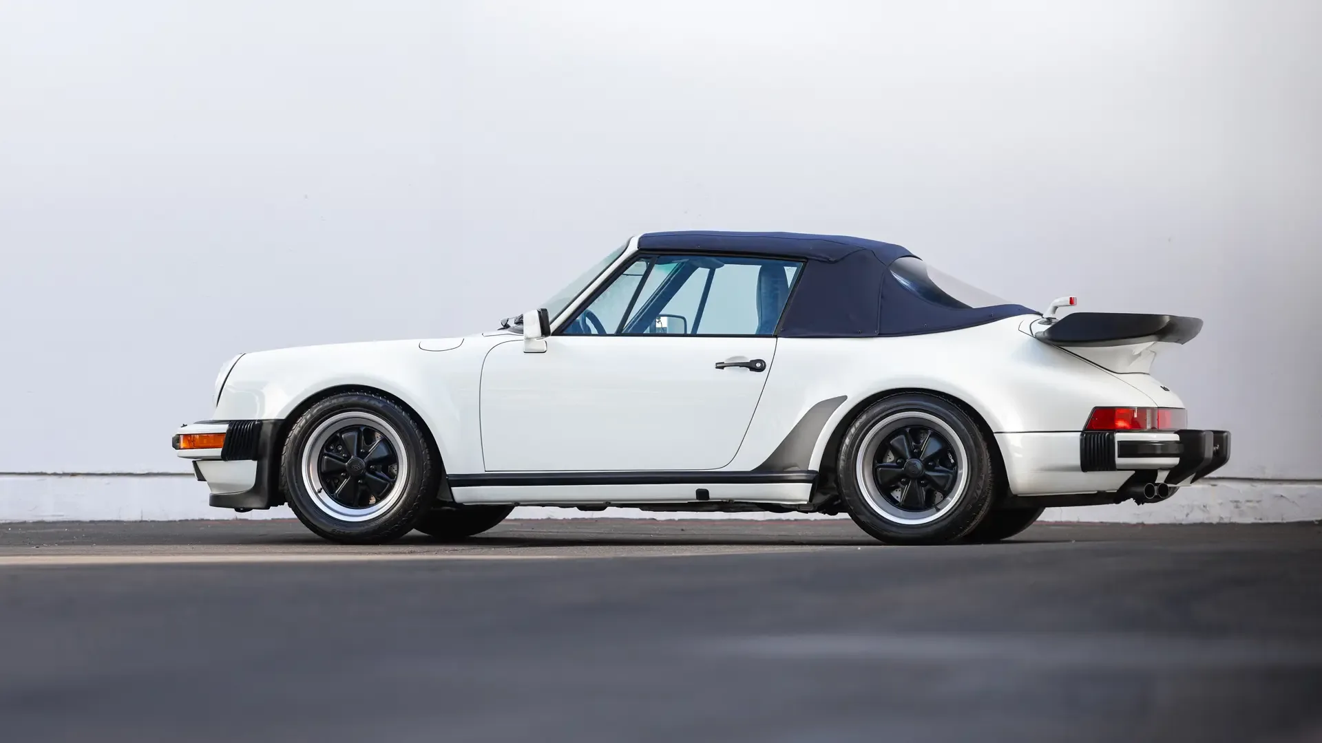 Seitenansicht eines weißen Porsche 911 Turbo Cabriolet (930) von 1989, mit dunkelblauem Stoffverdeck, auf Asphalt vor heller Wand.