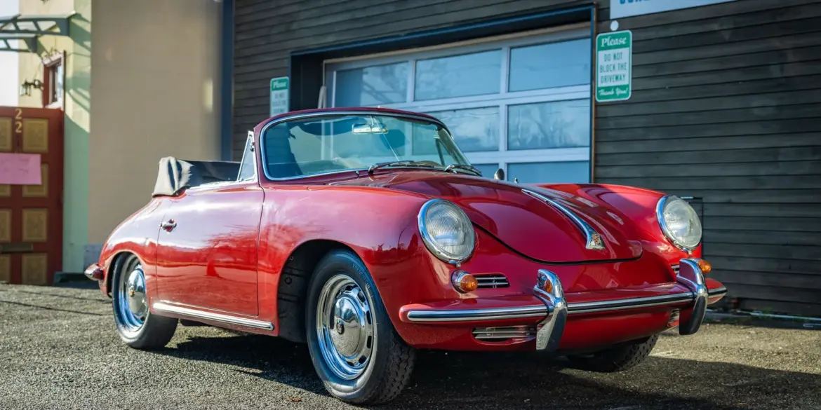 Rubyrotes Porsche 356C Cabriolet von 1965 mit geöffnetem Verdeck, seitlich und frontal fotografiert vor einer Garage.