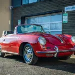 Rubyrotes Porsche 356C Cabriolet von 1965 mit geöffnetem Verdeck, seitlich und frontal fotografiert vor einer Garage.