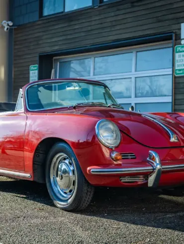 Rubyrotes Porsche 356C Cabriolet von 1965 mit geöffnetem Verdeck, seitlich und frontal fotografiert vor einer Garage.