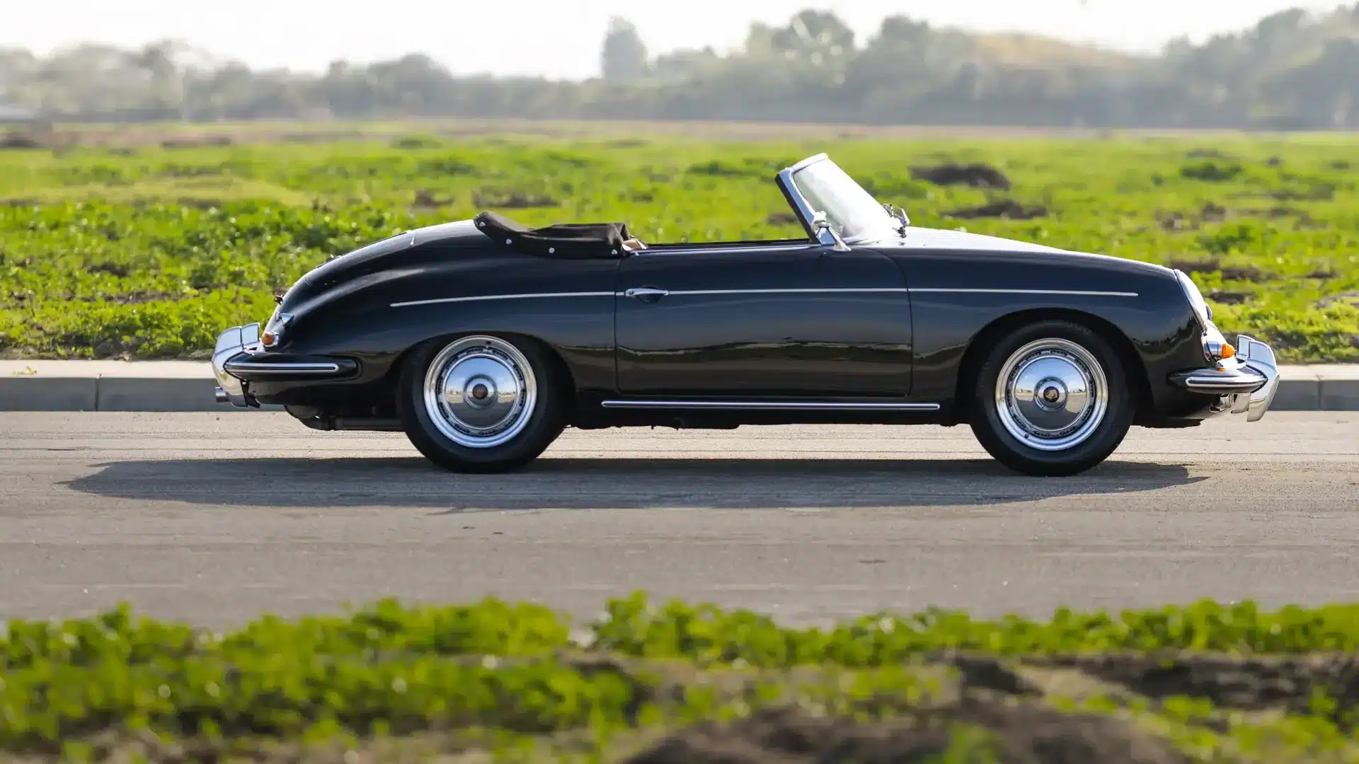 Schwarzer Porsche 356 B Super 90 Roadster von 1960 als Cabriolet, seitlich fotografiert auf einer Straße vor grüner Landschaft.