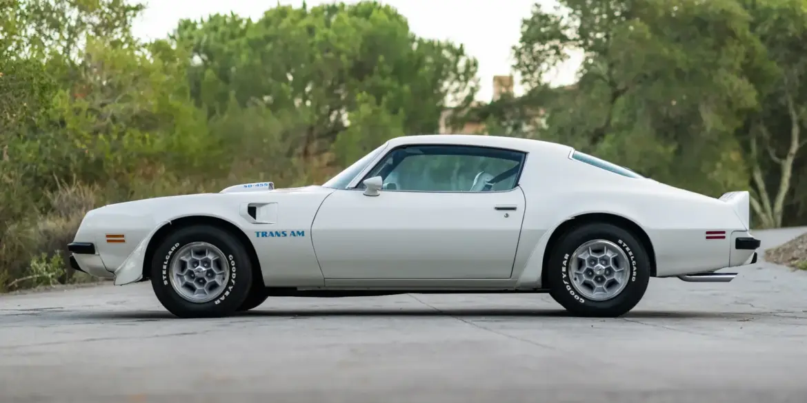Seitenansicht eines weißen Pontiac Trans Am SD-455 von 1974 mit blauer "Trans Am" Beschriftung und SD-455 Haube, stehend auf einer Straße im Grünen.