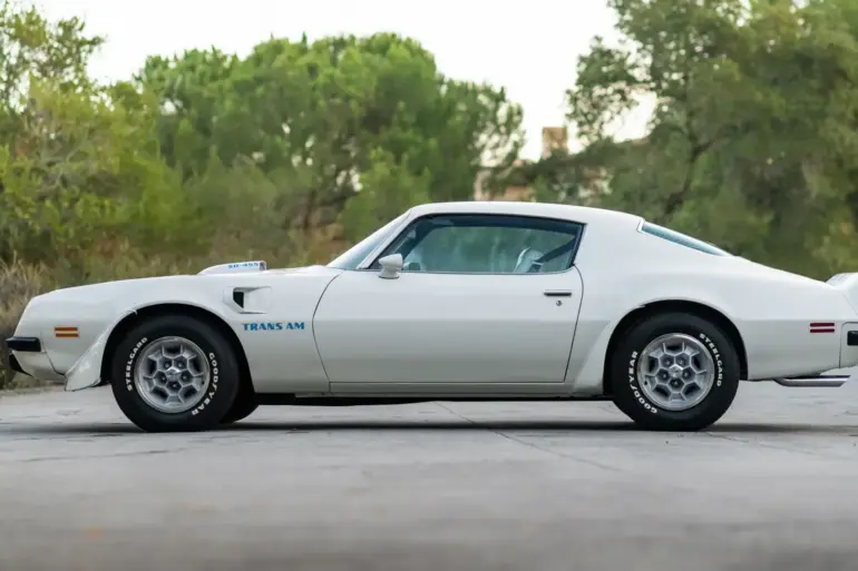 Seitenansicht eines weißen Pontiac Trans Am SD-455 von 1974 mit blauer "Trans Am" Beschriftung und SD-455 Haube, stehend auf einer Straße im Grünen.