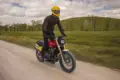 Fahrer auf einer Fantic Caballero 500 Scrambler Motorrad fährt auf einer Schotterstraße durch grüne Landschaft.