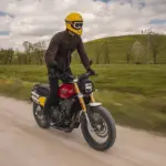 Fahrer auf einer Fantic Caballero 500 Scrambler Motorrad fährt auf einer Schotterstraße durch grüne Landschaft.