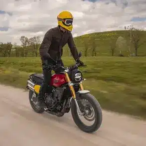Fahrer auf einer Fantic Caballero 500 Scrambler Motorrad fährt auf einer Schotterstraße durch grüne Landschaft.