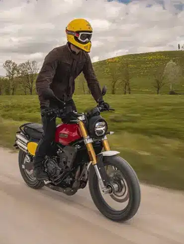 Fahrer auf einer Fantic Caballero 500 Scrambler Motorrad fährt auf einer Schotterstraße durch grüne Landschaft.