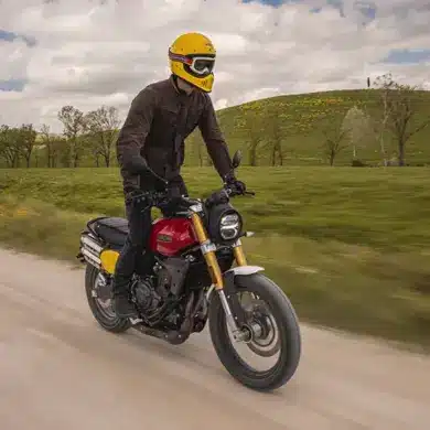 Fahrer auf einer Fantic Caballero 500 Scrambler Motorrad fährt auf einer Schotterstraße durch grüne Landschaft.