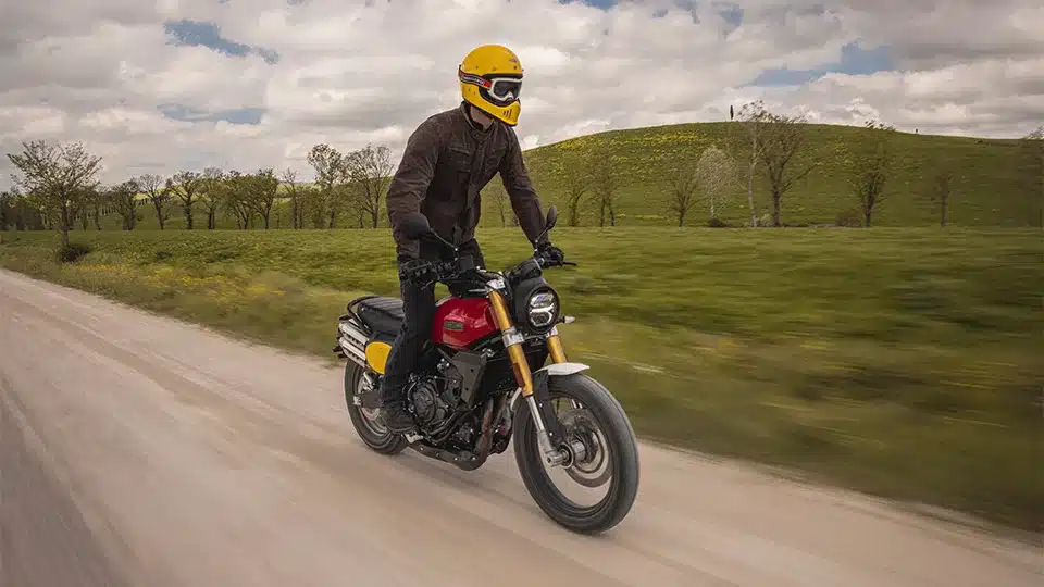 Fahrer auf einer Fantic Caballero 500 Scrambler Motorrad fährt auf einer Schotterstraße durch grüne Landschaft.