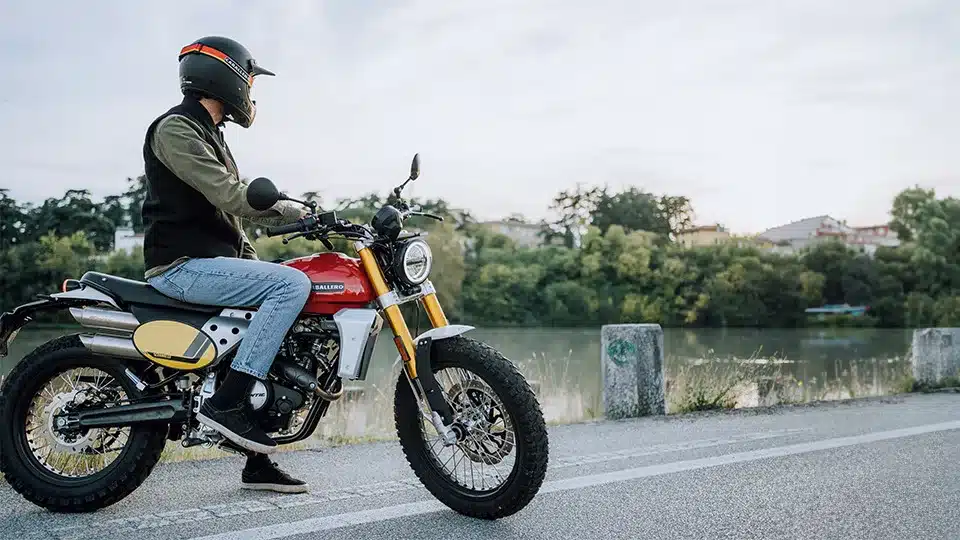 Fahrer sitzt auf einer Fantic Caballero 500 Scrambler mit rotem Tank, gelbem Seitendeckel und Doppelauspuffanlage am Flussufer.