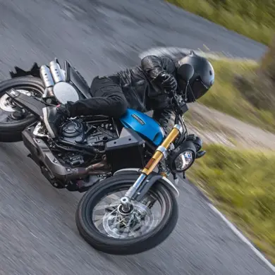Fantic Caballero 700 Scrambler-Motorrad in Blau auf einer kurvigen Straße, Fahrer mit schwarzer Ausrüstung.