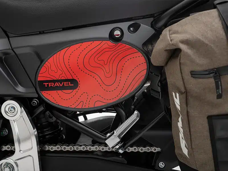 Rotes Seitenpanel mit 'Travel'-Schriftzug und Kartendesign an der Fantic Caballero Travel 700, daneben eine Packtasche am Motorrad.