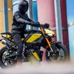 Fantic Stealth 125 Motorrad in Gelb, seitlich fotografiert, Fahrer mit kompletter Schutzkleidung, urbane Kulisse mit bunten Säulen.