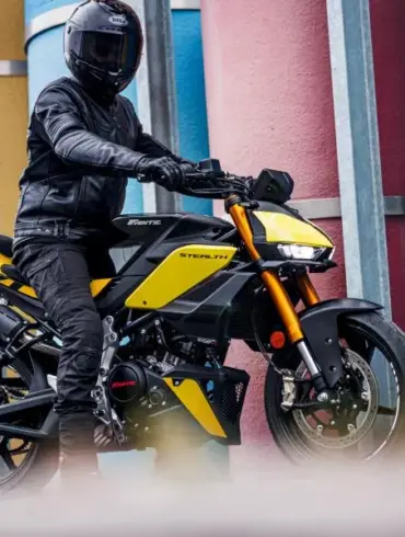 Fantic Stealth 125 Motorrad in Gelb, seitlich fotografiert, Fahrer mit kompletter Schutzkleidung, urbane Kulisse mit bunten Säulen.