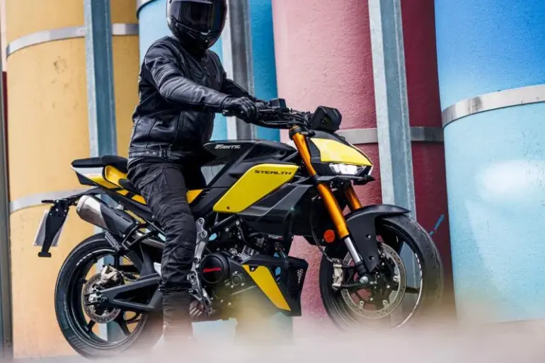 Fantic Stealth 125 Motorrad in Gelb, seitlich fotografiert, Fahrer mit kompletter Schutzkleidung, urbane Kulisse mit bunten Säulen.