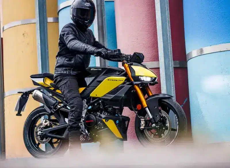 Fantic Stealth 125 Motorrad in Gelb, seitlich fotografiert, Fahrer mit kompletter Schutzkleidung, urbane Kulisse mit bunten Säulen.