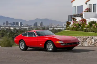 Ferrari 365 GTB/4 Daytona Berlinetta von 1970 in Rot, seitlich fotografiert vor einer Stadtansicht und einem modernen Haus.