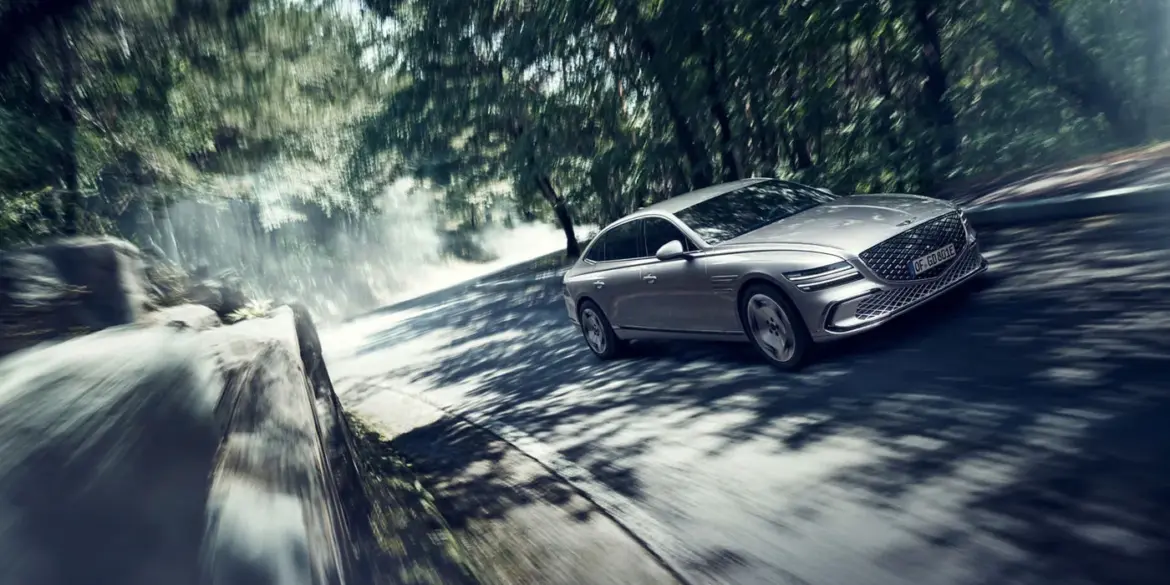 Genesis Electrified G80 als elektrische Limousine auf einer sonnigen, bewaldeten Straße in dynamischer Fahrt.