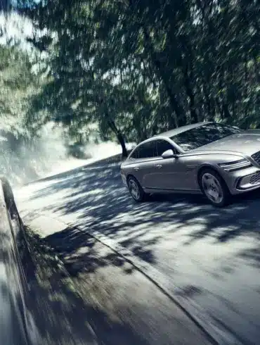 Genesis Electrified G80 als elektrische Limousine auf einer sonnigen, bewaldeten Straße in dynamischer Fahrt.