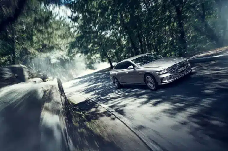 Genesis Electrified G80 als elektrische Limousine auf einer sonnigen, bewaldeten Straße in dynamischer Fahrt.