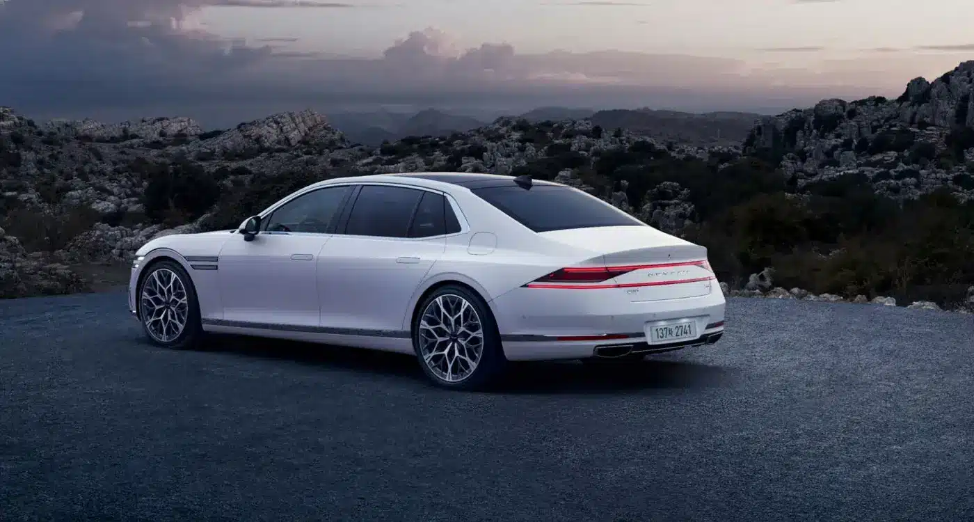 Weißer Genesis G90 von hinten auf einer Straße, umgeben von Bergen und bewölktem Himmel