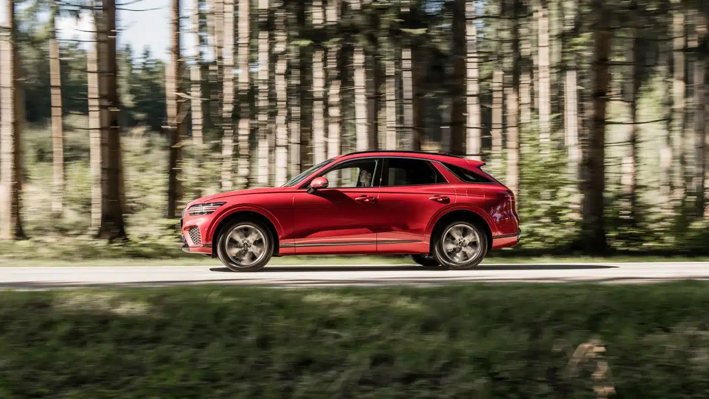 Genesis GV70 SUV in Rot fährt dynamisch auf einer Straße durch den Wald.