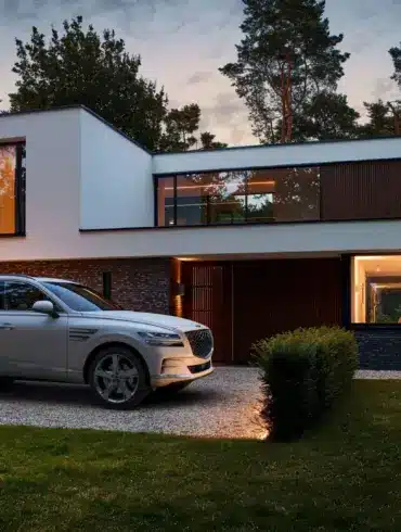 Ein silberner Genesis GV80 steht vor einem modernen Wohnhaus, im Hintergrund leuchtet warmes Licht aus den Fenstern.