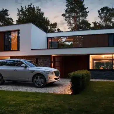 Ein silberner Genesis GV80 steht vor einem modernen Wohnhaus, im Hintergrund leuchtet warmes Licht aus den Fenstern.