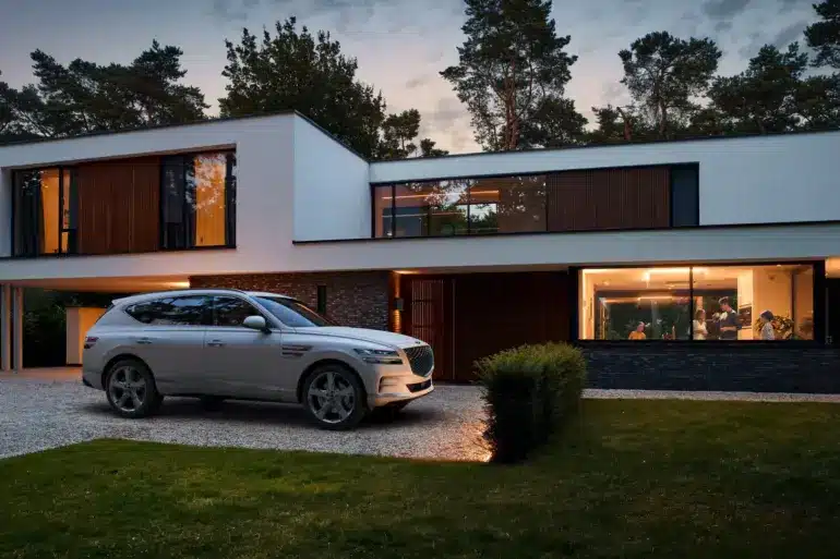 Ein silberner Genesis GV80 steht vor einem modernen Wohnhaus, im Hintergrund leuchtet warmes Licht aus den Fenstern.