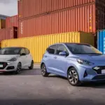 Zwei Hyundai i10 Modelle, einmal in Weiß und einmal in Hellblau, stehen nebeneinander auf einem Industriegelände vor bunten Containern.